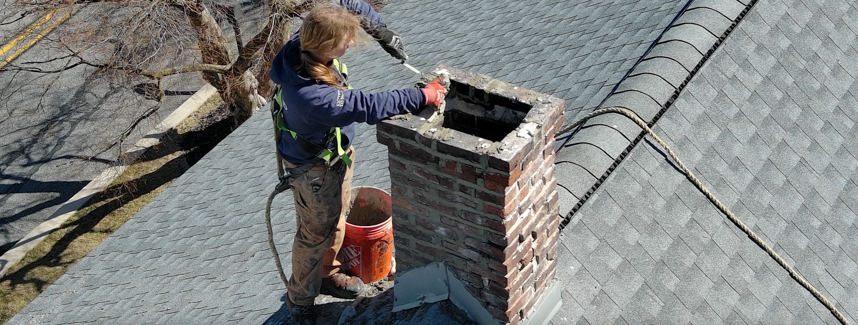 Person on a roof is working on a brick chimney. They are wearing safety gear.