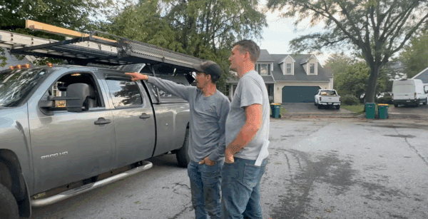 Two men near a work truck, one pointing upward. Suburban setting with houses and trees.
