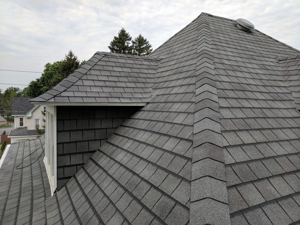 Gray asphalt shingle roof on a house, angled view. Overcast sky, trees and other houses visible.