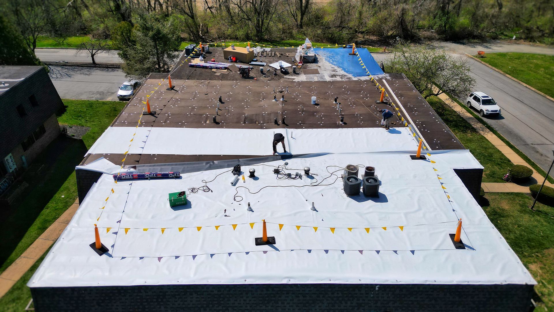 Workers on a flat roof replacing roofing material. Safety cones and debris scattered. Daytime, outdoors.
