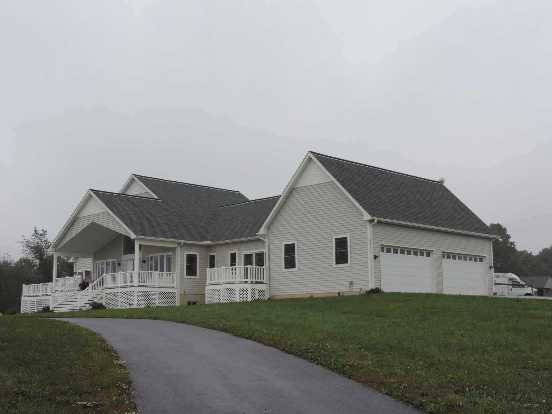 Light-colored house with attached garage, covered porch, and asphalt driveway on a grassy hill under a gray sky.