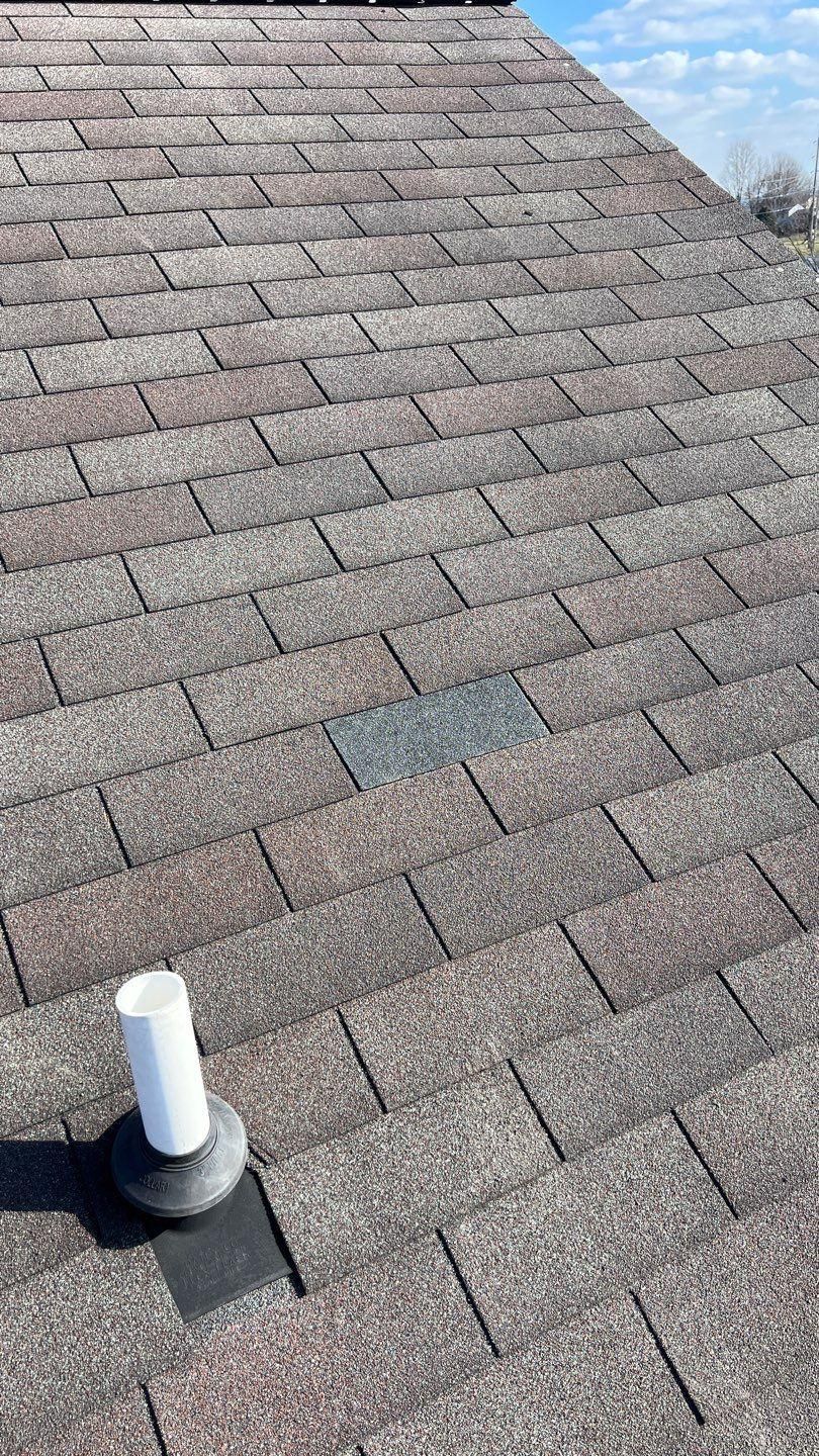 Close-up view of a shingled roof with a vent pipe and a dark area of missing or damaged shingles.