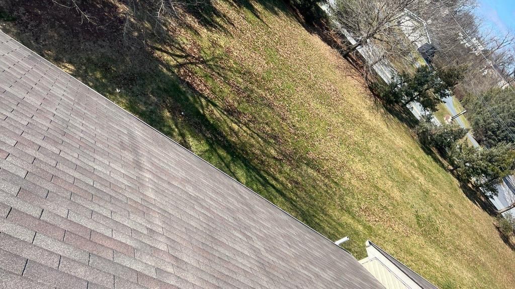 Rooftop view of grass, trees, and a street with buildings, likely residential. Shingles in the foreground.