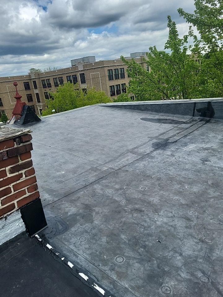 Flat, gray rooftop with brick chimney and dark trim, in an urban setting with apartment buildings.