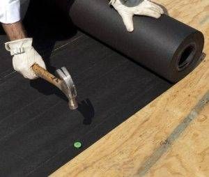 Person in gloves using a hammer to secure black roofing underlayment onto a wooden surface.