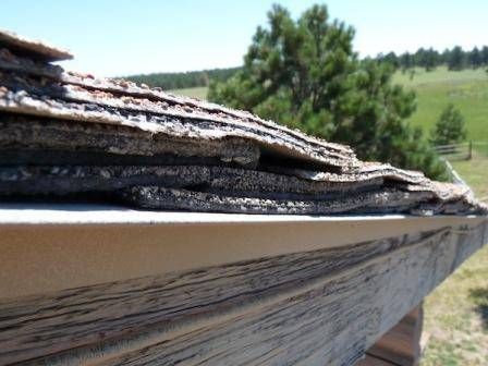 Close-up view of severely damaged asphalt shingles on a roof, with layers peeling away. Sunny outdoors.