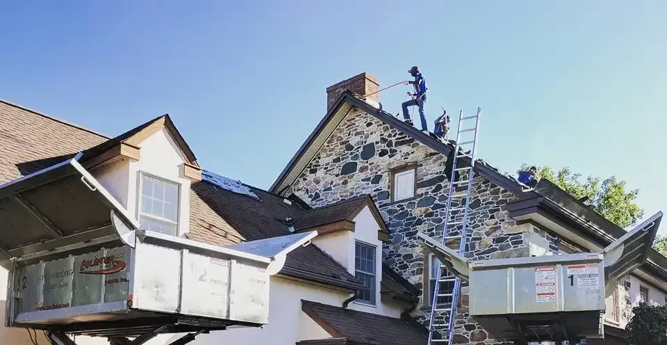 Workers on a roof, near a stone chimney, with a ladder and the blue sky above.