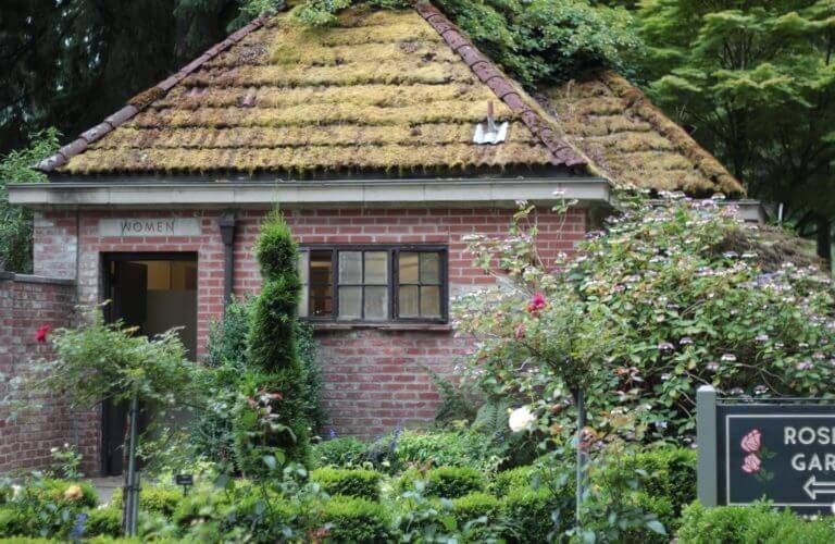 Brick building with mossy roof, surrounded by rose bushes in a garden. Sign reads