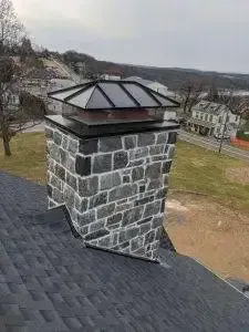Stone chimney with a glass top on a dark shingle roof, overlooking a road and houses.