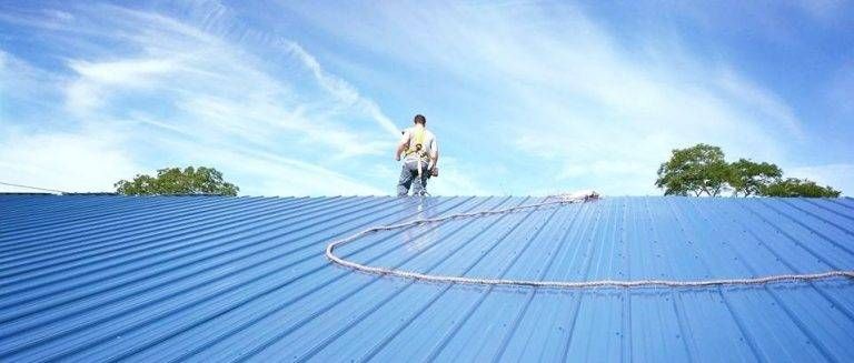 Man in safety harness working on a blue metal roof with a blue sky.