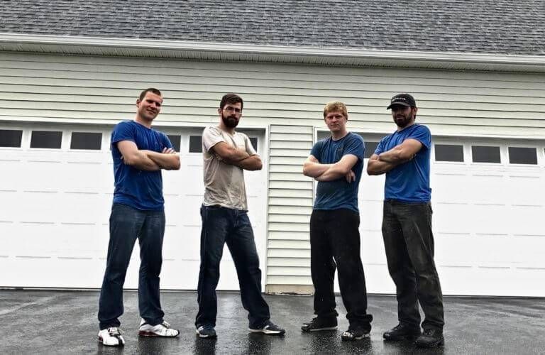 Four men stand arms crossed in front of a white garage door. They wear t-shirts and jeans or pants.