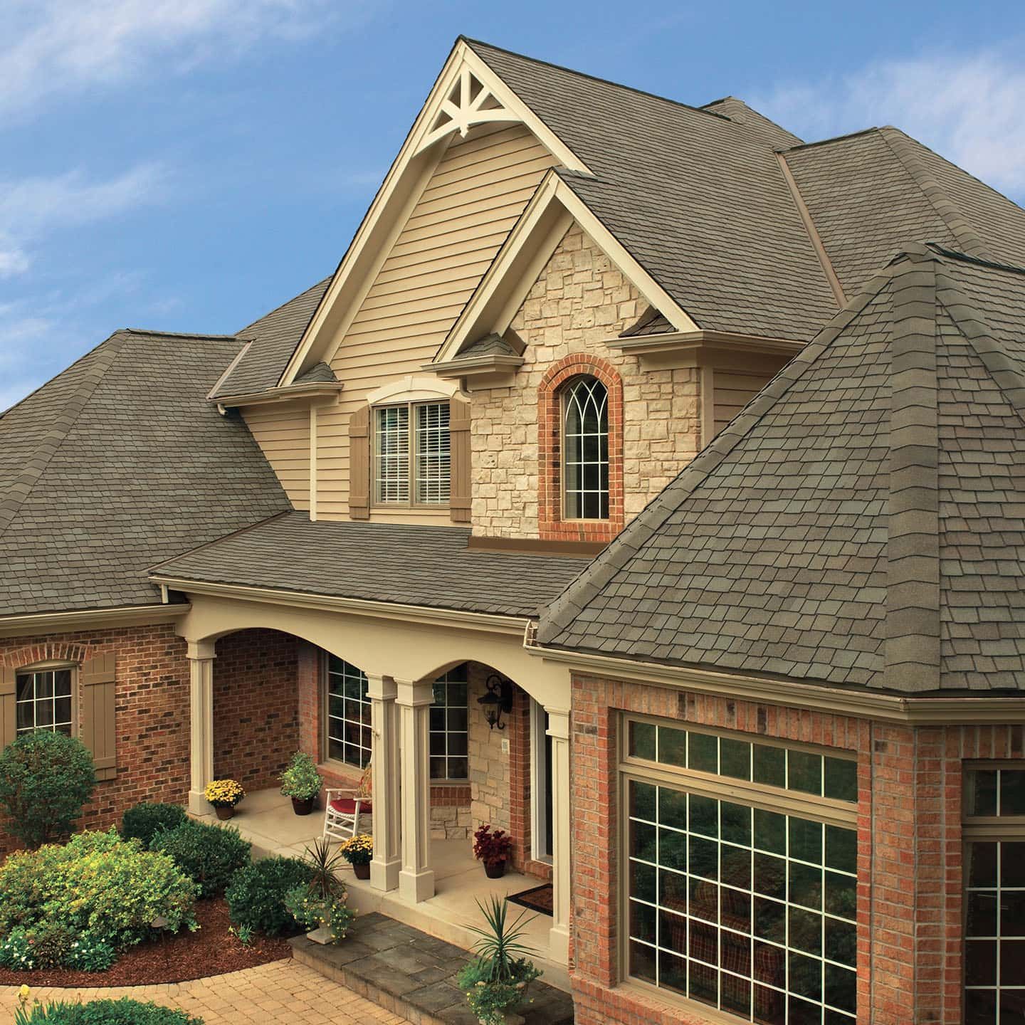 Two-story house with stone facade, beige siding, brown roof, and garage. Driveway and grass in front.