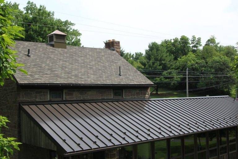Building with brown shingle roof and metal roof extension, surrounded by green trees.