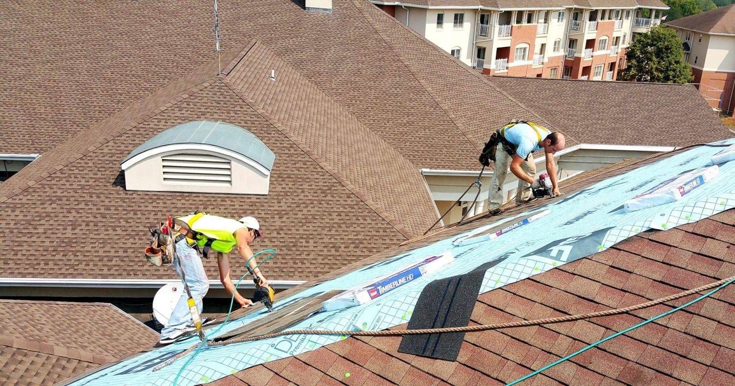 Two roofers installing shingles on a brown roof, wearing safety gear, with a multi-story building in the background.