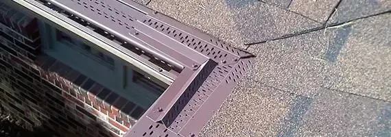 A roof with brown shingles, a brown gutter guard, and a brick wall.