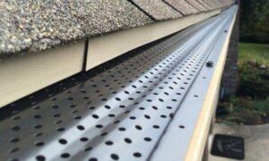 Close-up of a gutter with a perforated metal cover, installed on a roof with a tan fascia.