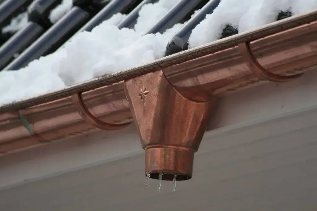 Copper gutter and downspout on a roof covered in snow; water drips from the downspout.