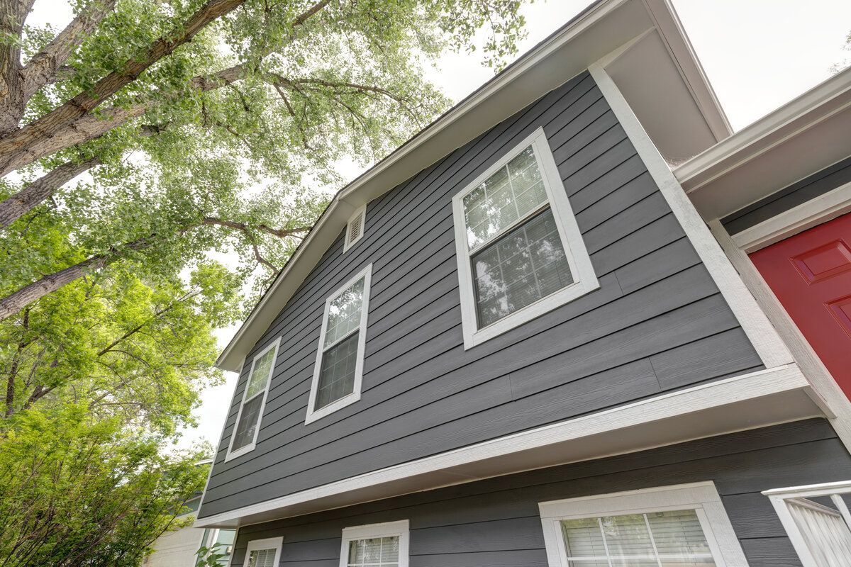 Gray house exterior with white-framed windows, red door, and surrounding tree branches against a cloudy sky.