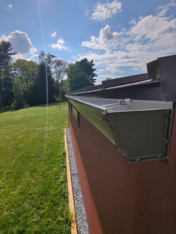 A metal gutter installed along the edge of a brown, stucco-style building next to a grassy lawn under a blue sky.
