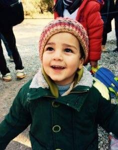 Smiling child wearing a knitted hat and green jacket; outdoors, other people in the background.