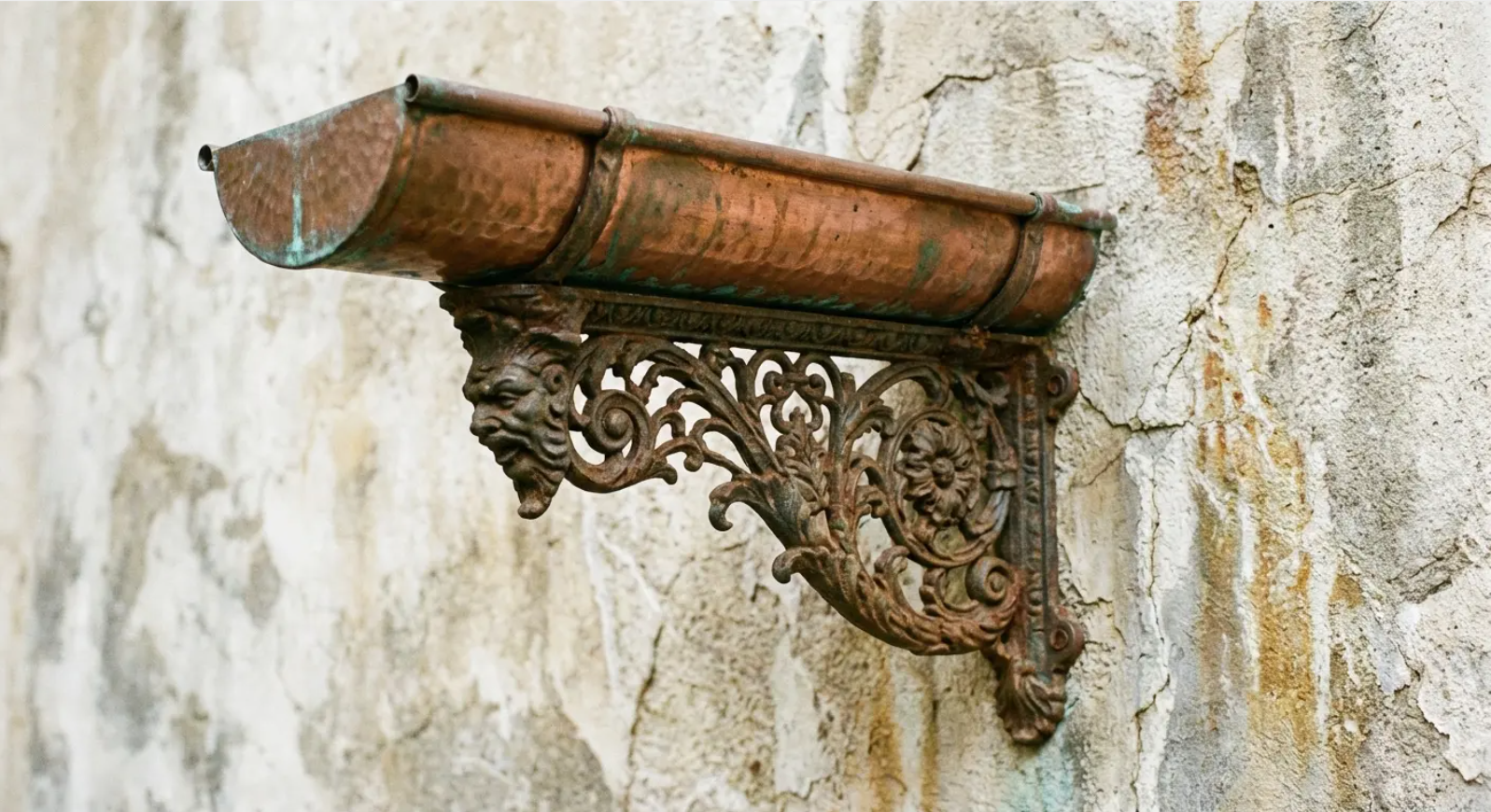 Rusty copper gutter with ornate bracket on weathered stucco wall.