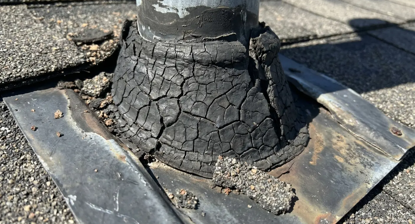 Cracked black rubber roof boot around a metal pipe, on a shingle roof.