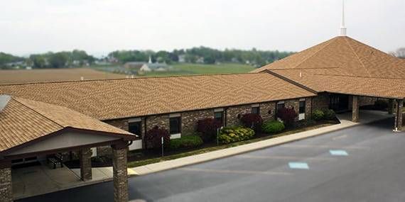 A one-story brick building with a brown roof and a covered entrance in front of a field and small town.