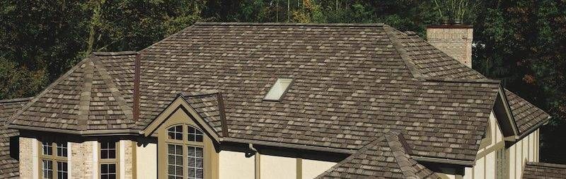 Brown-tiled roof of a house with multiple gables, chimney, and a small skylight. Trees in the background.