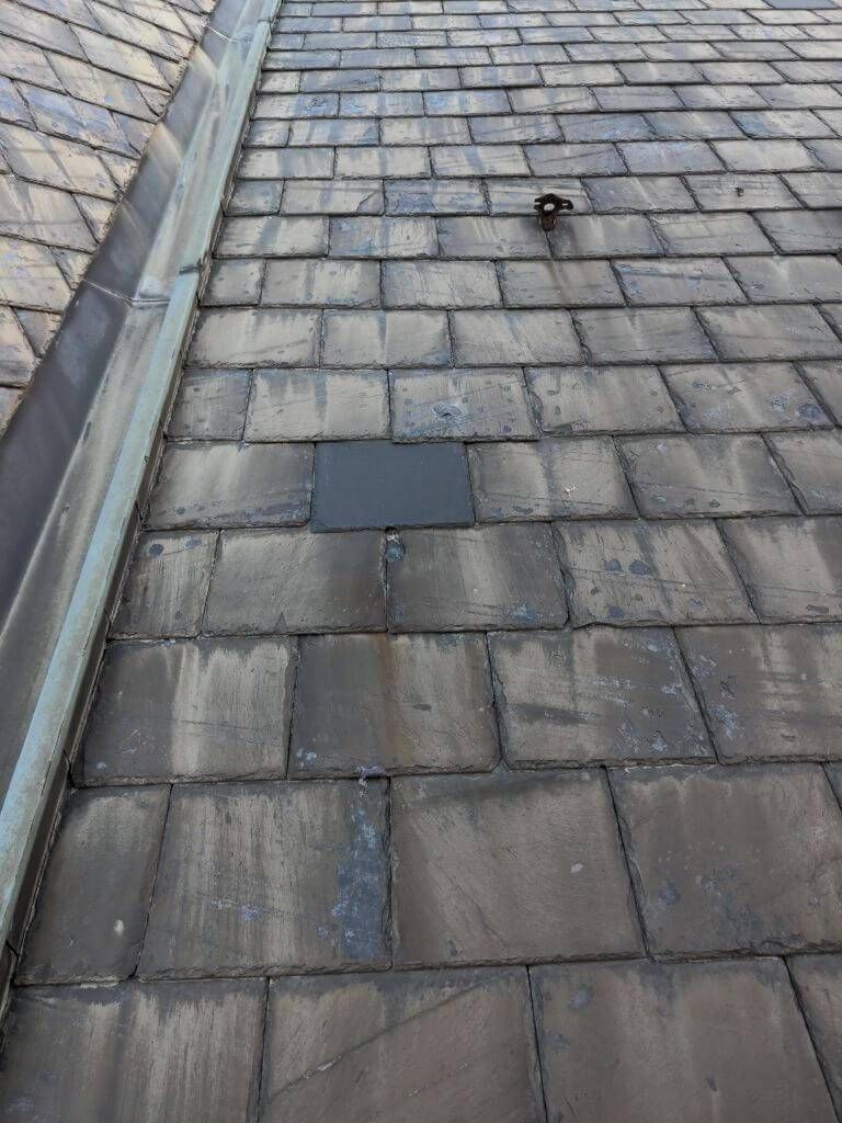 Dark slate roof with a single, dark grey replacement tile, next to a gutter.