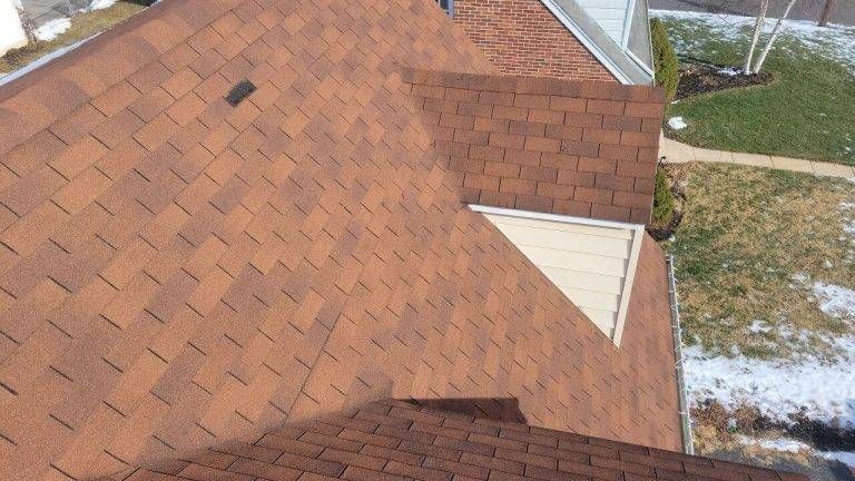 Brown shingle roof of a house with a white triangular window and partial snow on the ground.