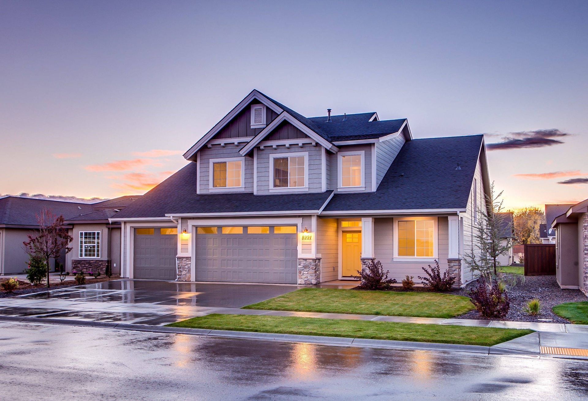 Two-story house with gray siding, two-car garage, and green lawn on a wet street at dusk.