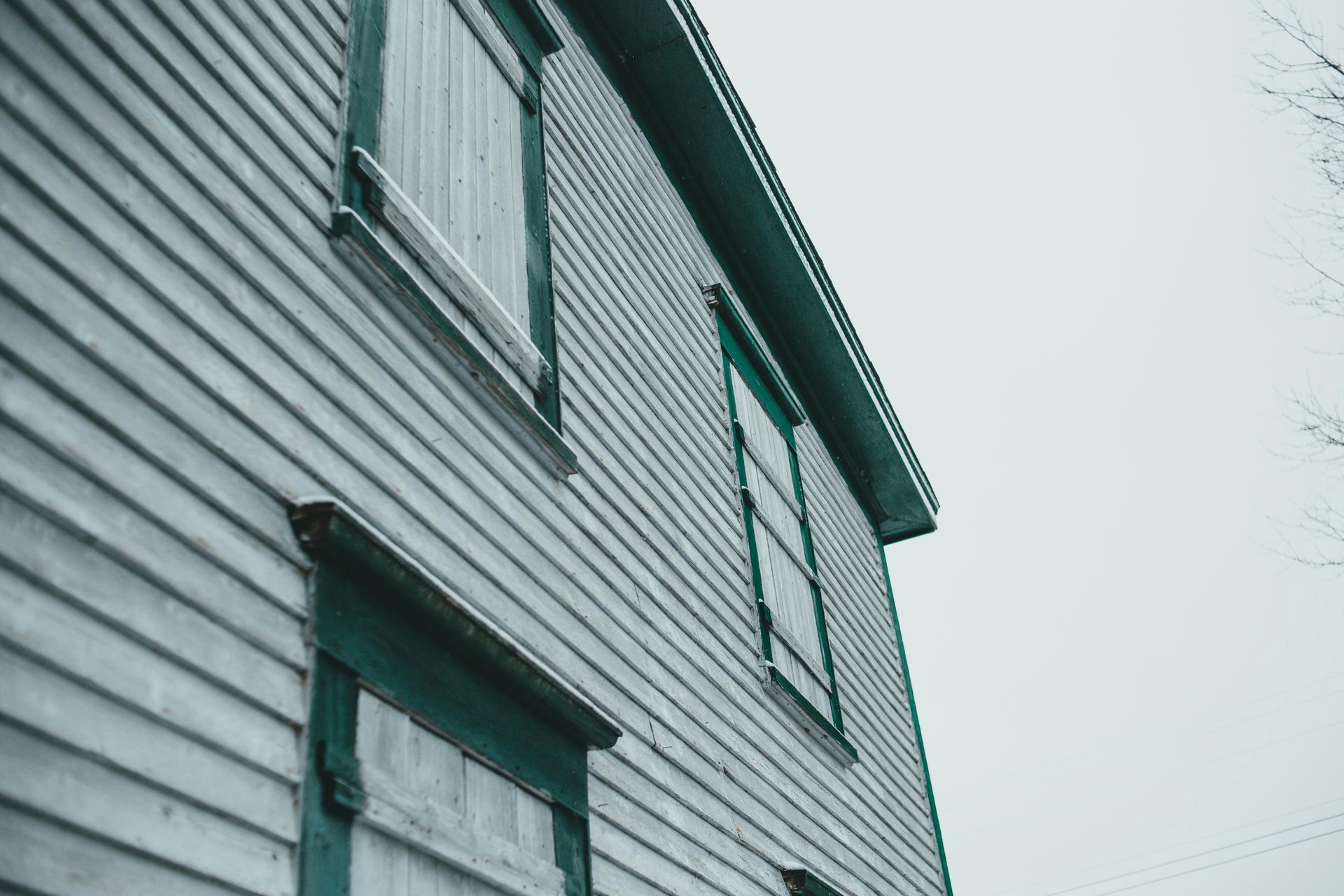 Side view of a weathered, light blue house with two boarded windows and a dark teal trim against a gray sky.