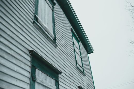 Side view of a weathered, light blue house with two boarded windows and a dark teal trim against a gray sky.