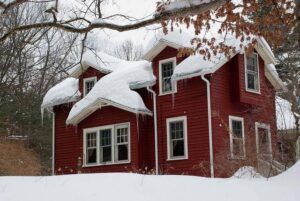 Red house in winter, snow-covered roof and windows, icicles, bare trees, and snowy ground.