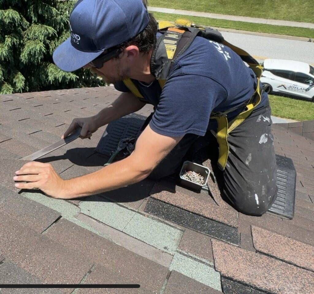 Roofer kneels on a shingled roof, cutting shingles with a utility knife, wearing a safety harness and cap.