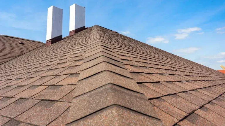 Brown shingled roof with two white chimneys against a blue sky.