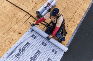 Roofer rolling out a gray underlayment on a wooden roof. Rolls of material and equipment are nearby.