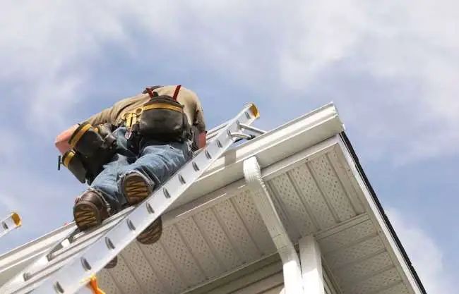 Person climbing a ladder to access a roof with a gutter, blue sky in the background.