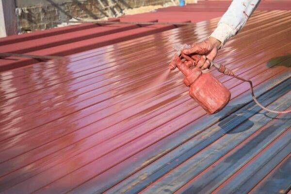 A person spraying red paint onto a corrugated metal roof.