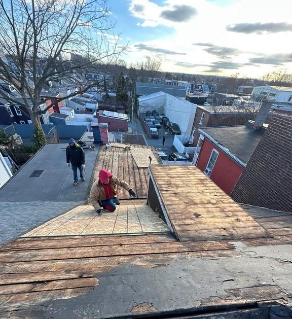 Two people on a rooftop, one kneeling inspecting old wood. Buildings and a street are in the background.