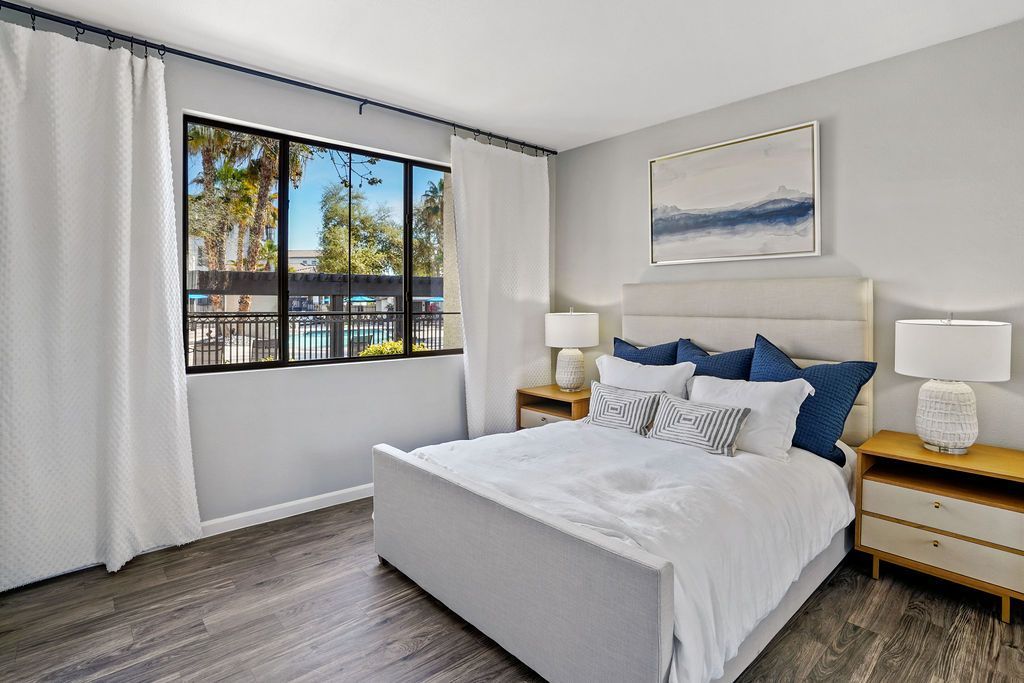 Bedroom in an apartment with a queen bed, two nightstands, lamps, and a large window with white curtains.