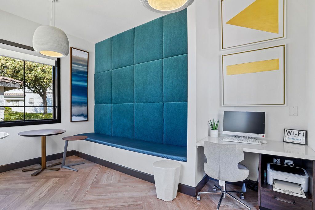 Sunlit community lounge with a blue padded wall, window seating, and a desk with a computer.