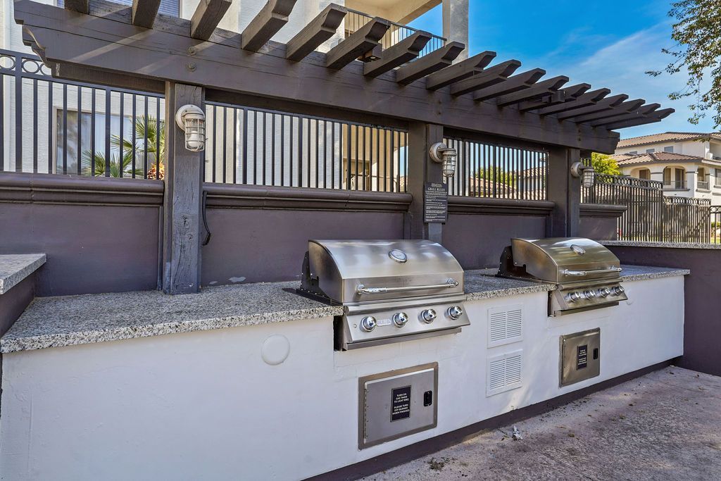 Outdoor communal grilling area with two stainless steel BBQ grills on a granite counter under a pergola.