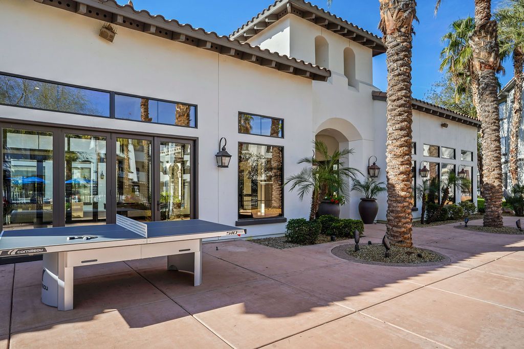 Exterior of a white apartment building with large glass doors, palm trees, and a ping-pong table in a sunny courtyard.