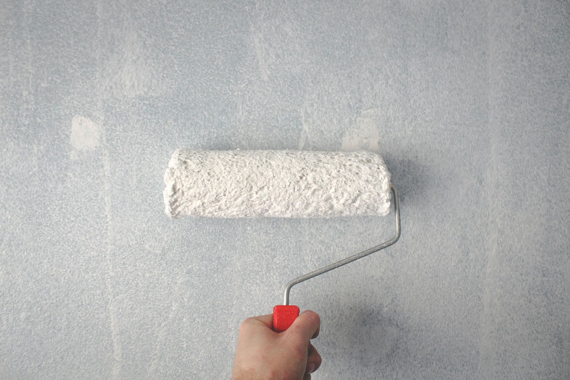 Hand holding a paint roller applying white paint to a light blue wall.