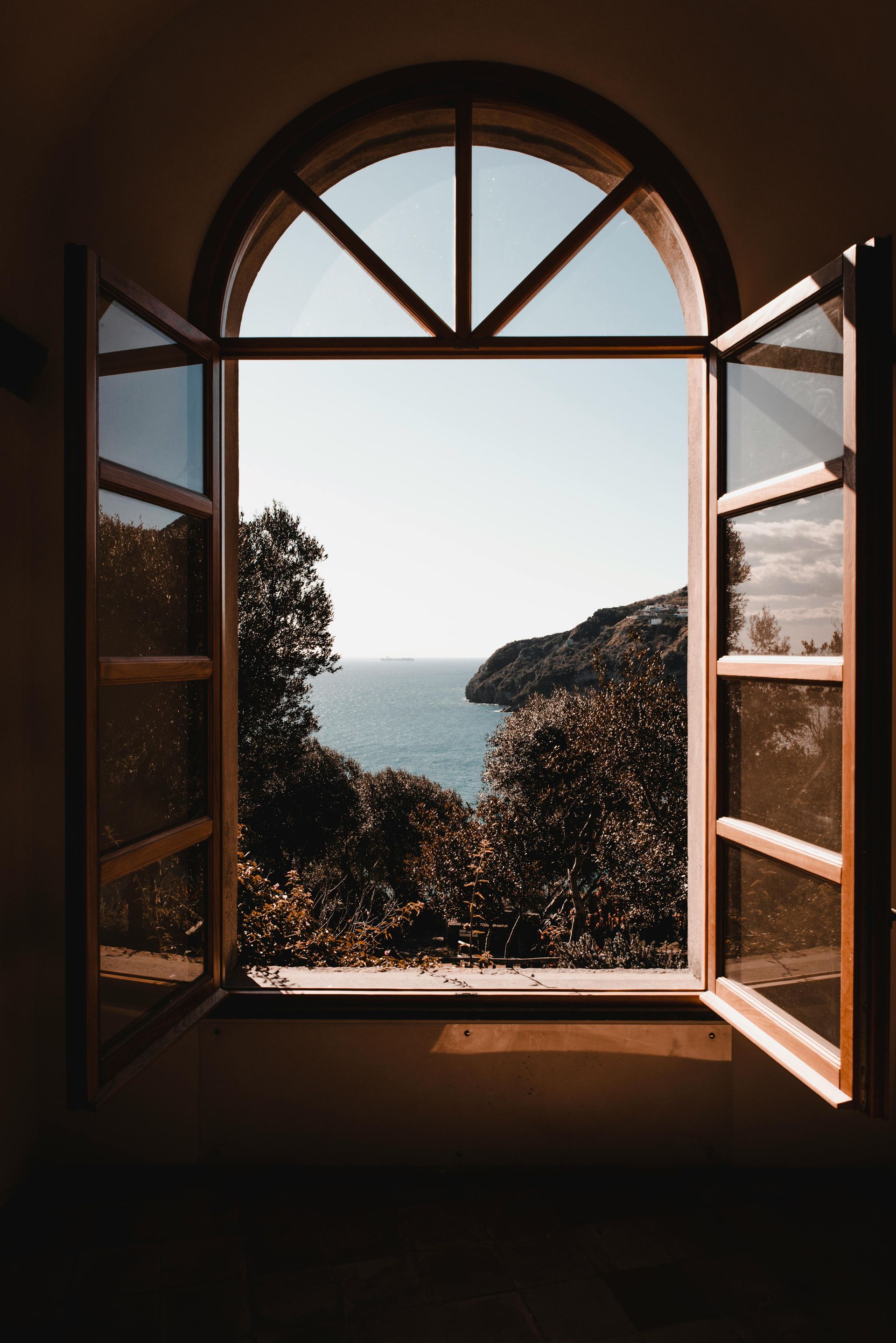 Open arched window framing a view of the sea and green-covered cliffs under a bright sky.