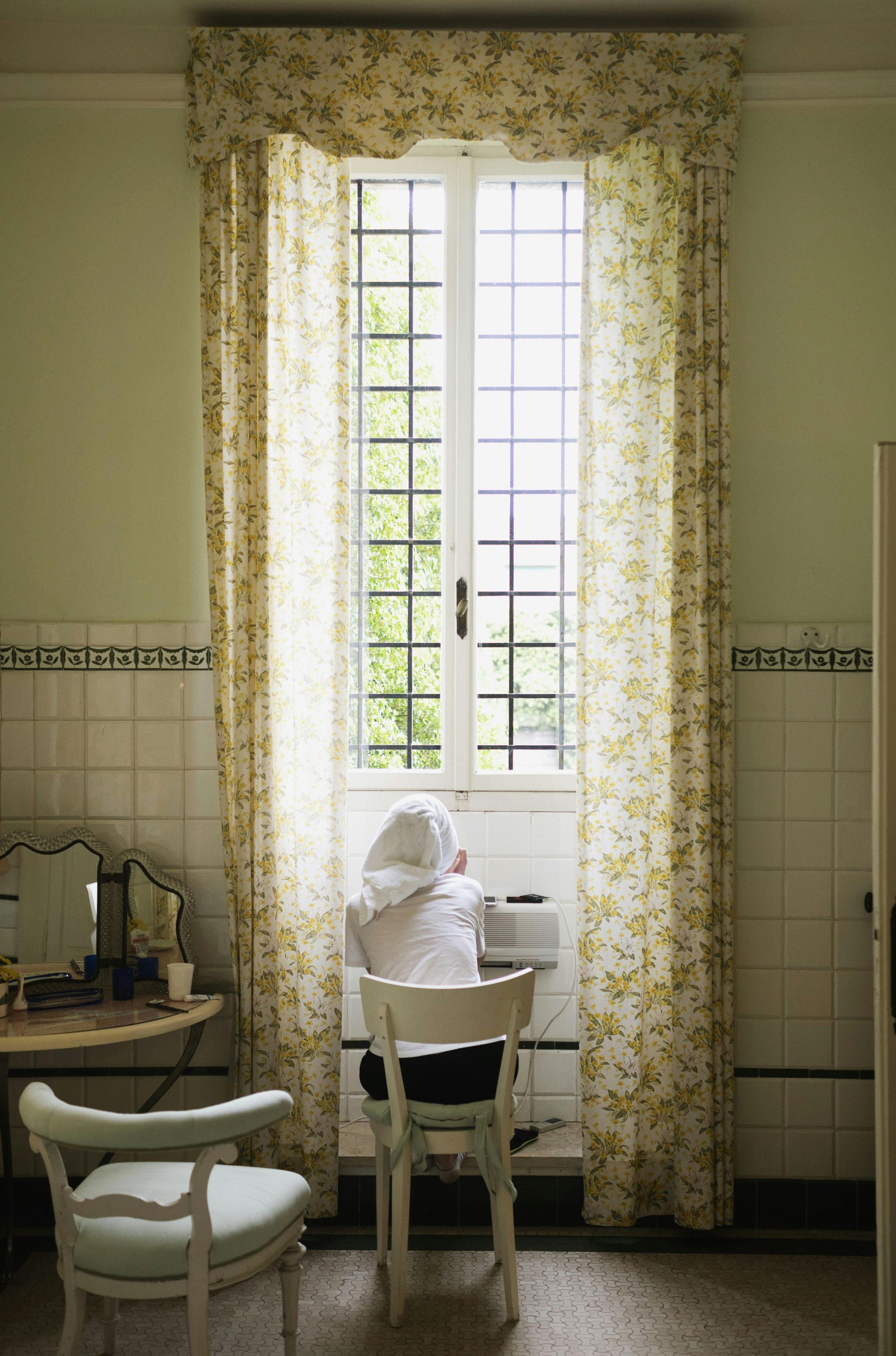 Person looking out a tall window with floral curtains in a bathroom setting.