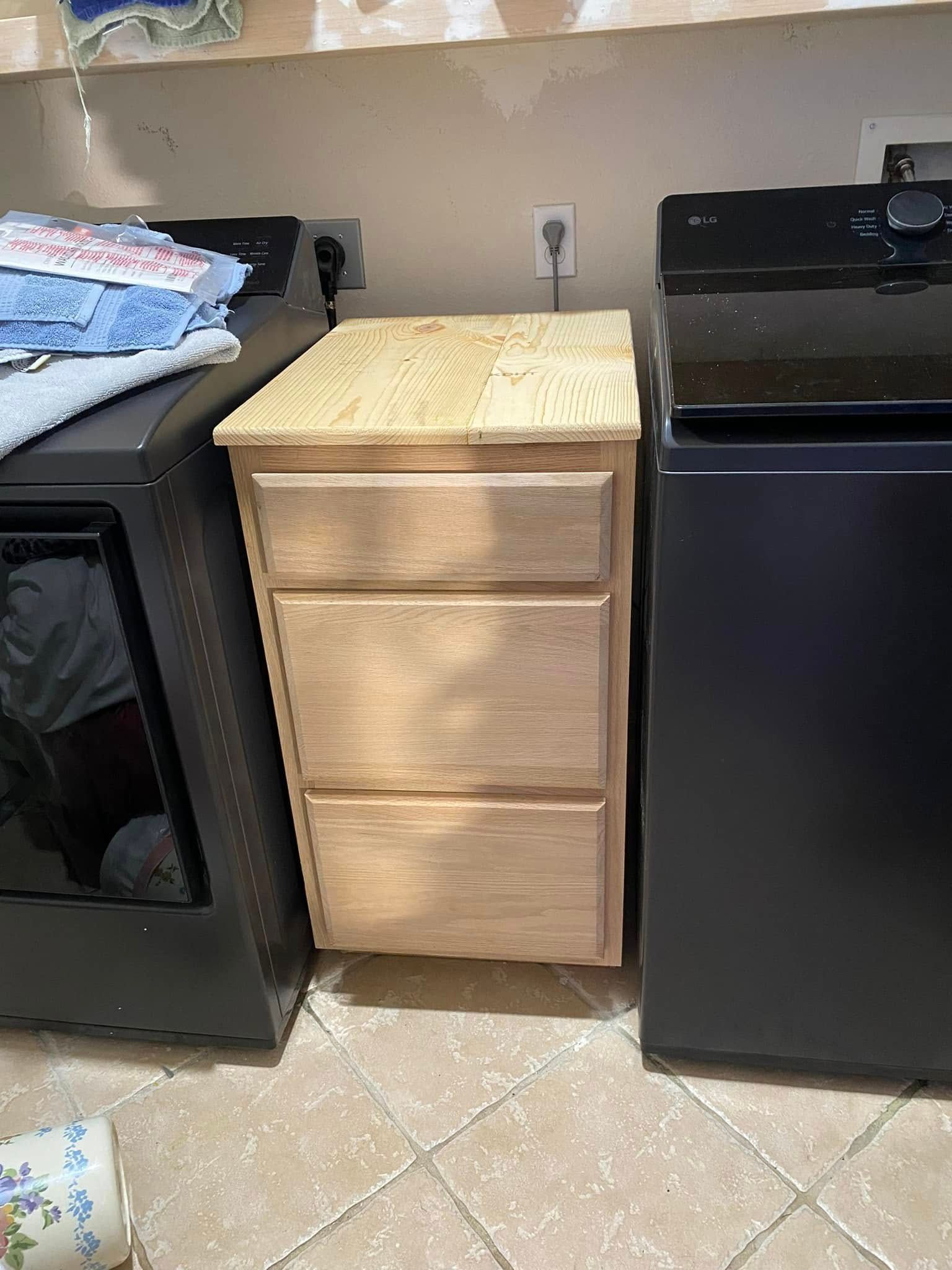 Wooden cabinet with three drawers sits between a black washing machine and a dryer in a laundry room.