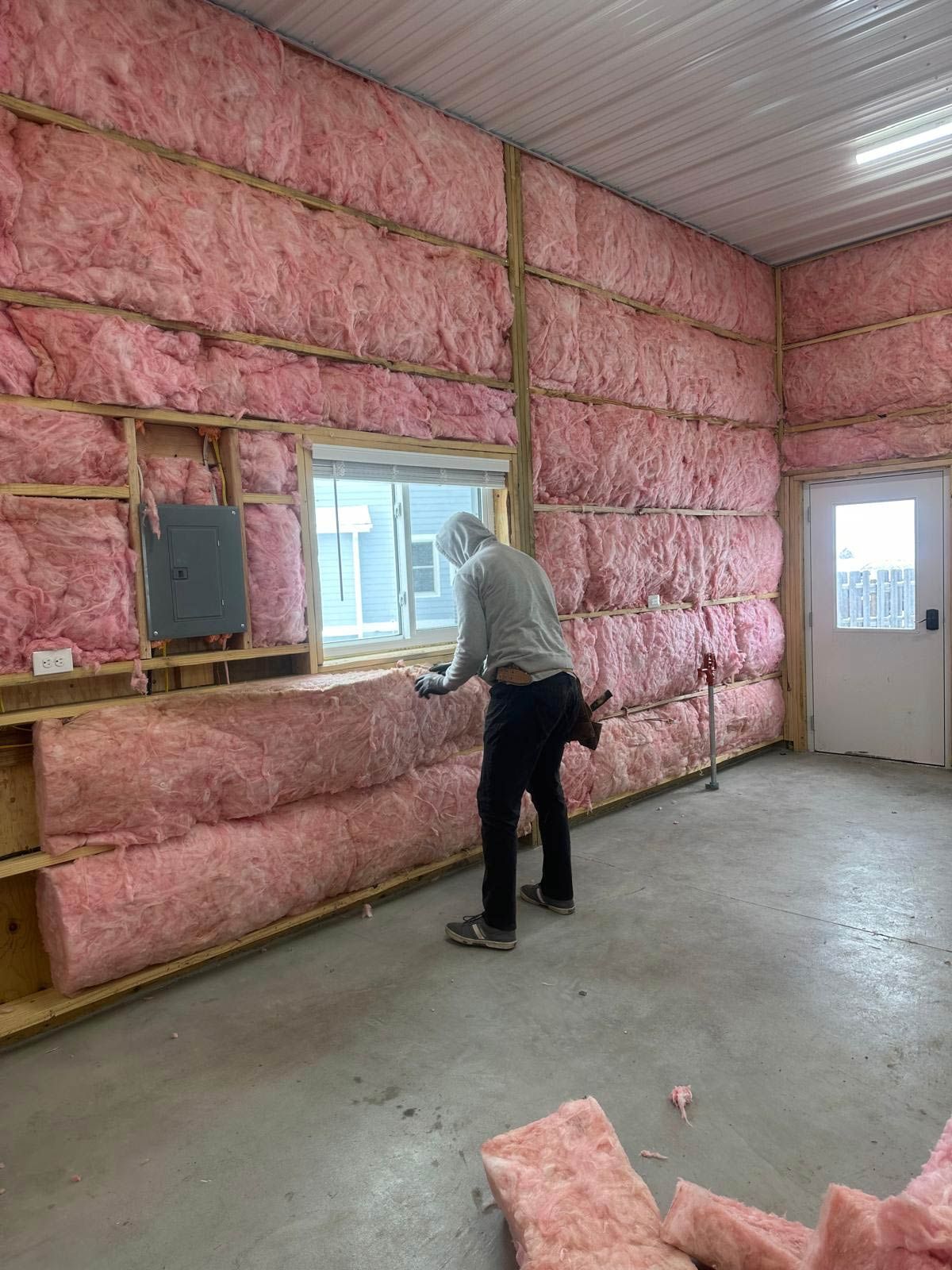 Worker installing pink insulation in a building's wall, near a window and electrical box.