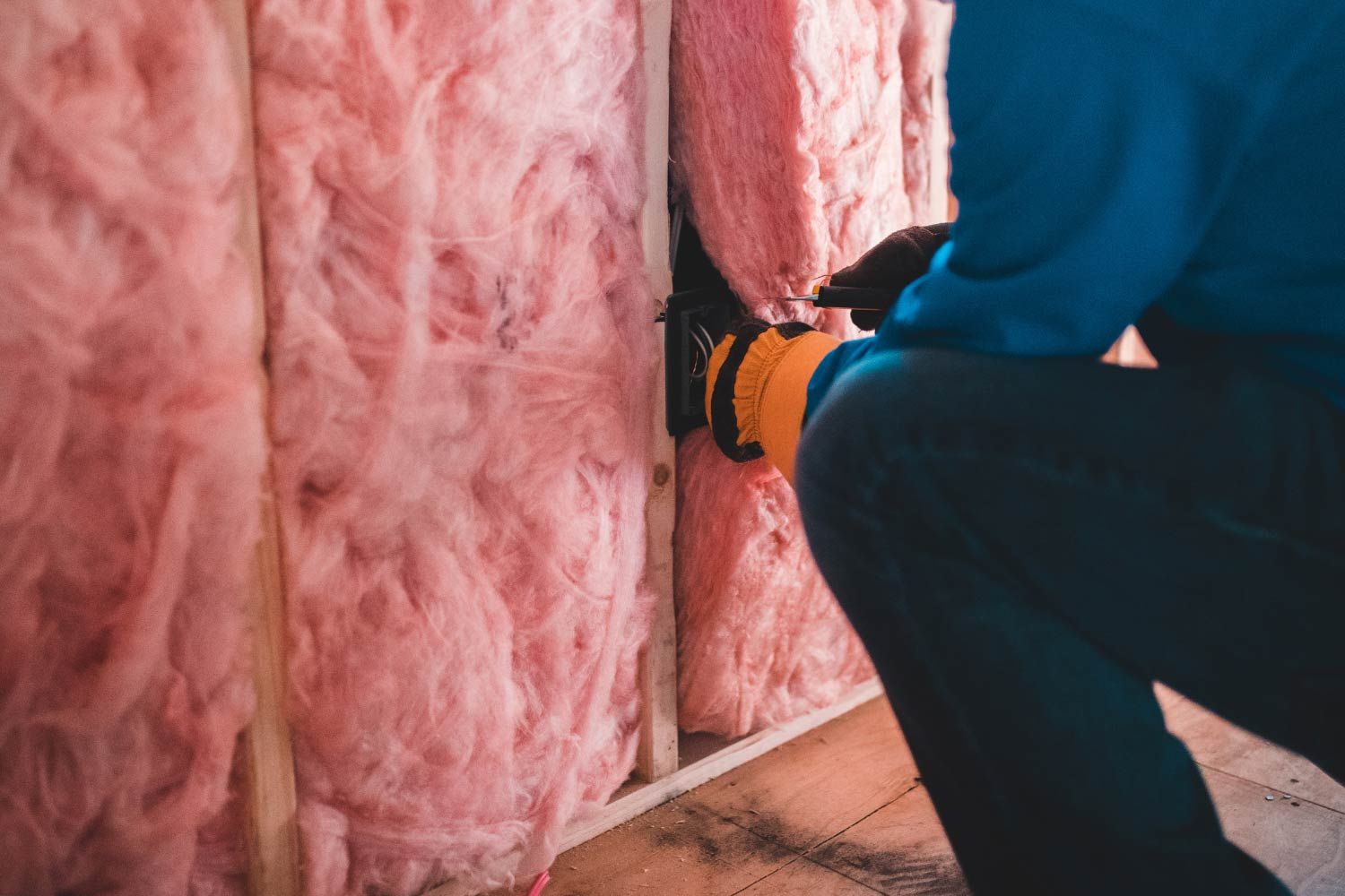 Person installs pink fiberglass insulation between wooden wall studs.